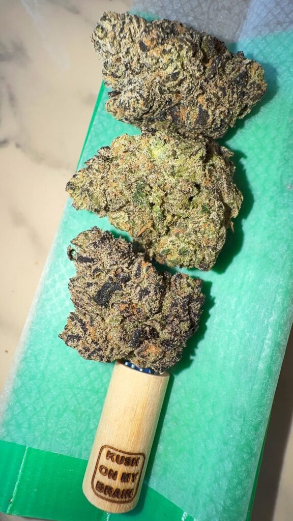 Macro view of a premium Melon Berries cannabis bud showing its unique spiky structure and dark purple hues, featured alongside a Kapow Berries 7g jar, a black metal grinder, and Bitcoin coins on a luxury desk setup for a connoisseur review.